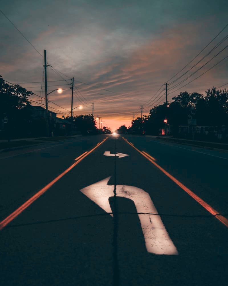 A quiet suburban road at dusk with two faded white arrows painted on the pavement, one pointing straight and one curving left