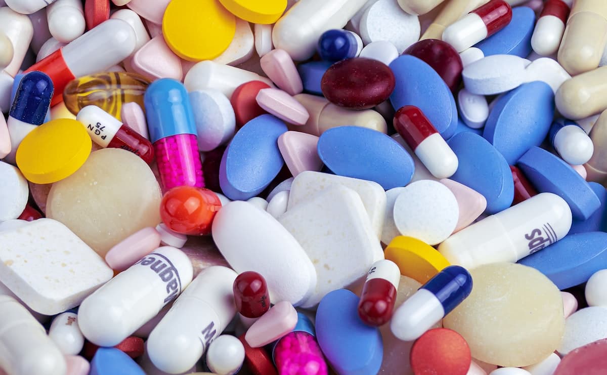 A white supplement bottle with a white cap beside a small pile of white capsules on a teal background
