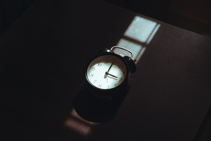 A black analog alarm clock on a dark surface lit by a sliver of window light at night
