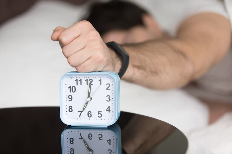 A hand pressing down on a light-blue alarm clock beside a person lying in bed under white sheets