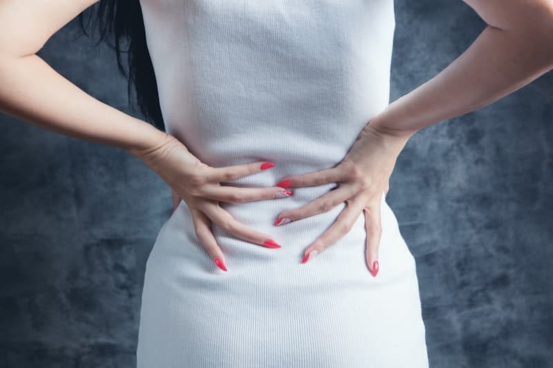 Woman in a white top holding her lower abdomen with red-painted nails, suggesting digestive discomfort
